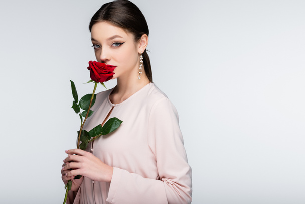 brunette young woman in earrings and blouse smelling red rose isolated on grey - Foto, Imagem