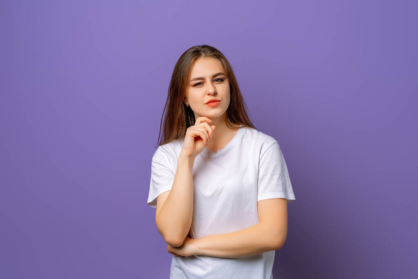 Retrato de una chica morena pensativa que parece impresionada con incredulidad con la mano en la barbilla, de pie en blanco camiseta blanca sobre fondo púrpura - Foto, Imagen