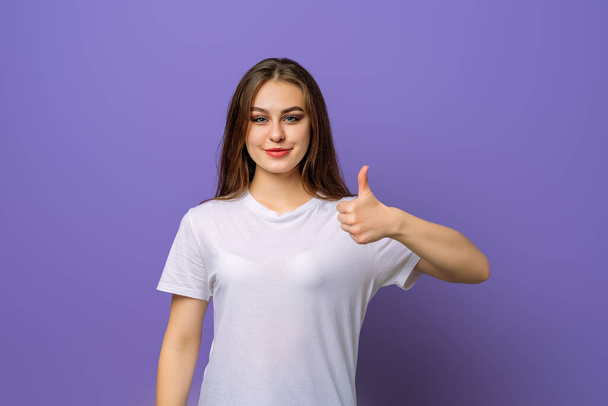 Gelukkig glimlachend mooie jonge brunette vrouw tonen duim omhoog gebaar, staande over paarse achtergrond. Positieve emoties concept - Foto, afbeelding