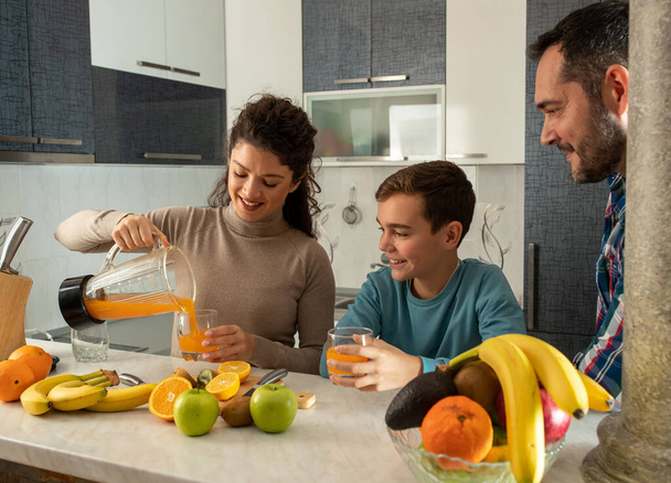 Mother pouring fresh frutty juice from blender to son and husband in kitchen at home - Foto, imagen