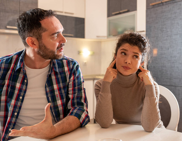 Husband and wife having argument at dining table at home. Man explaining something with dominant posture and woman holding fingers in ears and having bored expression - Foto, Imagem
