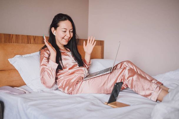 Young happy brunette woman with wireless earphones lying in bed in pink silk pajamas with smartphone and laptop computer, have online video chat. Blogger broadcasts live morning stream - Fotó, kép