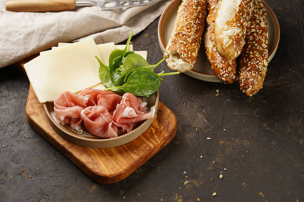 Savoury breakfast ingredients: brown lye bread sticks garnished with oats , pork slices prosciutto, hard cheese gouda and fresh greens on wooden boards - Foto, imagen