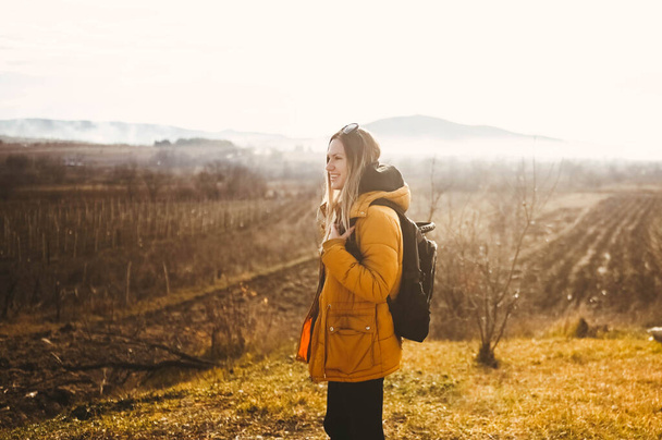 Young happy backpacker woman tourist traveling alone and posing at dawn with fog on background of beautiful nature with mountains in autumn. Blonde smiling lady in yellow down jacket. - Foto, imagen