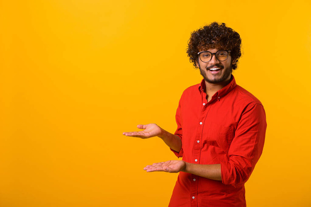 Good looking joyful man with beard presenting advertising area on his palm and pointing to copy space, holding empty place for commercial. Indoor studio shot isolated on orange - Zdjęcie, obraz