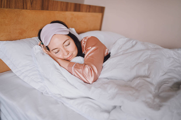 Ελκυστική χαμογελαστή νεαρή γυναίκα με ροζ μεταξωτές πιτζάμες και μάσκα ύπνου ματιών που απλώνεται στο κρεβάτι ξυπνώντας μόνη χαρούμενη έννοια, ξύπνια μετά από υγιή ύπνο σε άνετο κρεβάτι και στρώμα απολαύστε καλημέρα. - Φωτογραφία, εικόνα