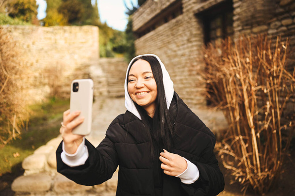 Young happy woman tourist traveling alone and making selfie with attractions by smartphone, have video chat. Blogger broadcasts live stream. Brunette lady in black down jacket and white hoodie - Фото, изображение