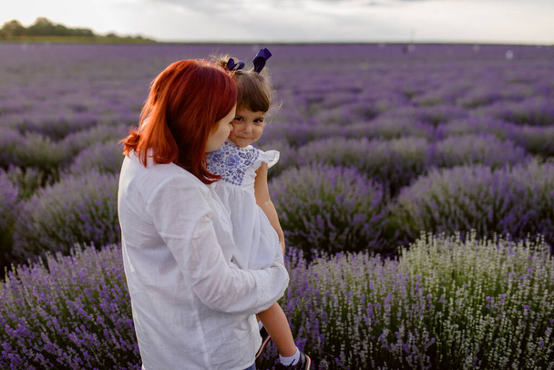 Young happy family concept. Mother is kissing her daughter in the middle of a lavender field. They all are wearing white clothes. Beautiful lavender flower - Fotoğraf, Görsel