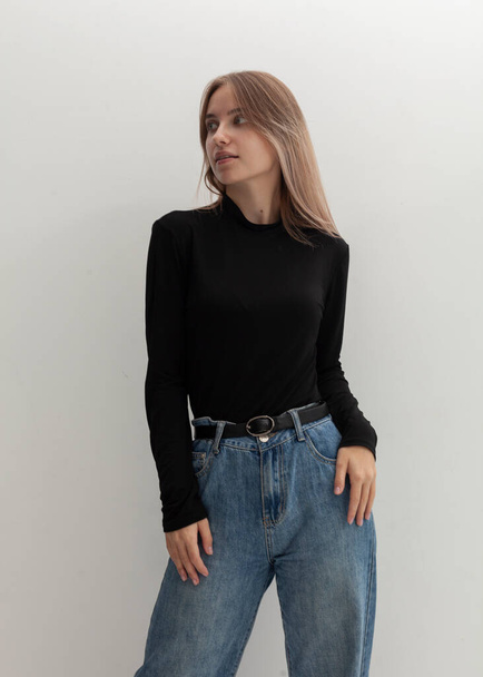 portrait of a beautiful blonde in a black turtleneck and jeans on a light background - Photo, Image