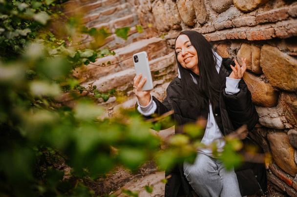 Young happy woman tourist traveling alone and making selfie with attractions by smartphone, have video chat. Blogger broadcasts live stream. Brunette lady in black down jacket and white hoodie - Foto, Imagem