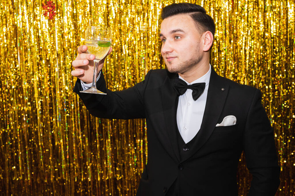 Stylový mladý muž v černém obleku s motýlem u zlaté stěny drží sklenici nápoje a zvedá ji, slavnostní náladu, usmívá se a dívá se stranou. Dovolená, párty. Selektivní zaměření - Fotografie, Obrázek