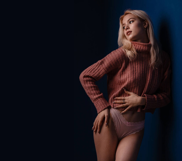 Gorgeous woman posing in sweater and sexy pink panties. Advertising underwear sensual slim fit body on dark blue background free space for text. Slim woman in sexy erotic underwear warm tone. - Φωτογραφία, εικόνα