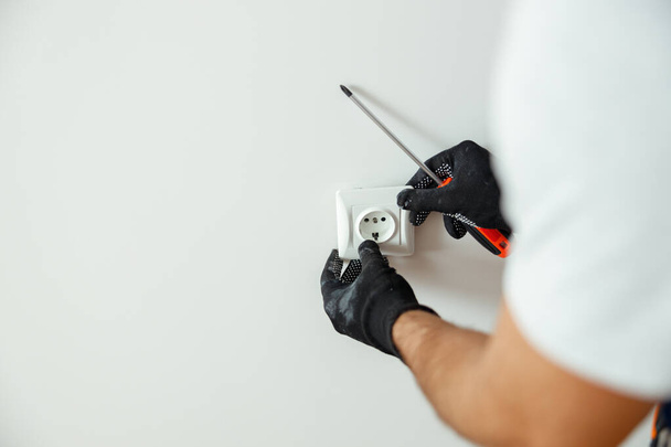 Close up of hands of male worker, professional electrician in protective gloves using screwdriver while installing new electrical socket outlet after renovation work - Foto, afbeelding