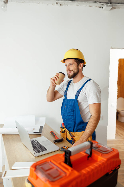 Ontspannen klusjesman in uniform en harde hoed drinken van koffie tijdens een pauze tijdens het doen van renovatiewerkzaamheden binnen - Foto, afbeelding