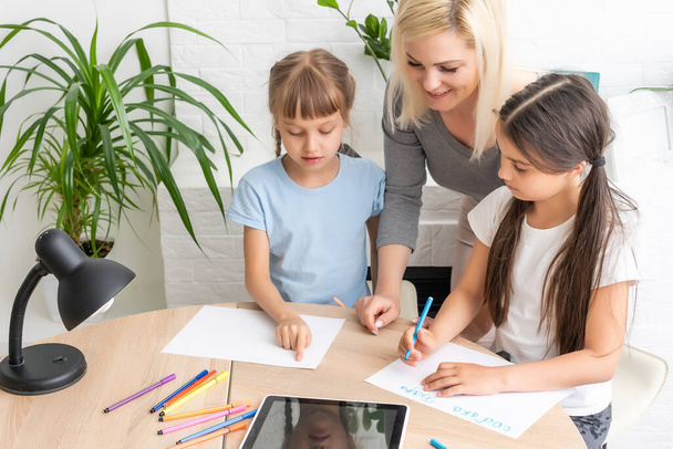 Korepetytorka lub matka zastępcza pomaga uroczej dziewczynie z białej szkoły odrabiać lekcje siedząc przy stole. Różnorodna niania i dziecko uczą się pisania w notatniku studiując w domu - Zdjęcie, obraz