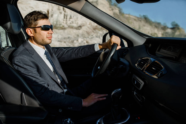 Молодий чоловік бізнесмен в куртці, сорочці, краватці і сонцезахисних окулярах водить машину
 - Фото, зображення