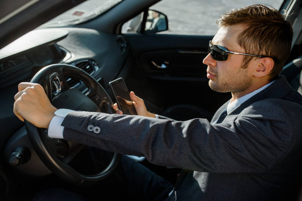 Молодий чоловік бізнесмен в куртці, сорочці, краватці і сонцезахисних окулярах водить машину
 - Фото, зображення