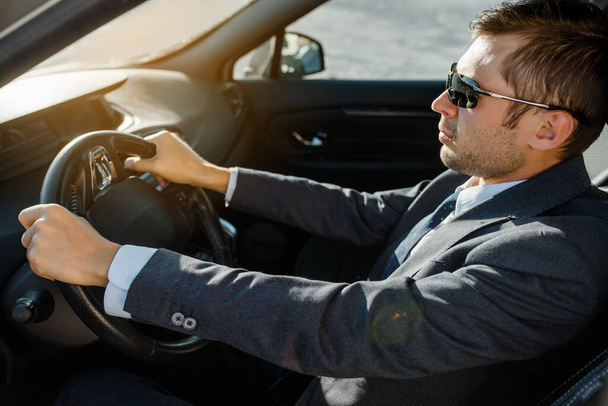 Νεαρός επιχειρηματίας με σακάκι, πουκάμισο, γραβάτα και γυαλιά ηλίου οδηγεί αυτοκίνητο. - Φωτογραφία, εικόνα