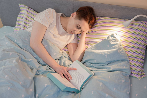 Young red-haired woman fell asleep in bed while reading before bedtime. Woman fell asleep over an open book in the light of the bedside lamp. - Фото, зображення