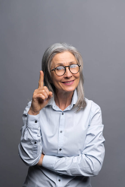Foto vertical de la mujer de negocios madura de pelo gris sonriente aislada sobre fondo gris señala con el dedo hacia arriba. Dama prestando atención - Foto, Imagen