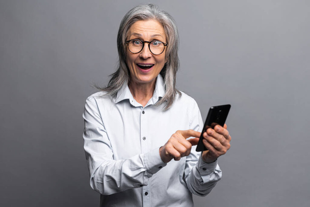 Surprised business woman in formal shirt pointing finger at smartphone while feeling shocked with new phone features. Indoor studio shot isolated on grey - Fotografie, Obrázek