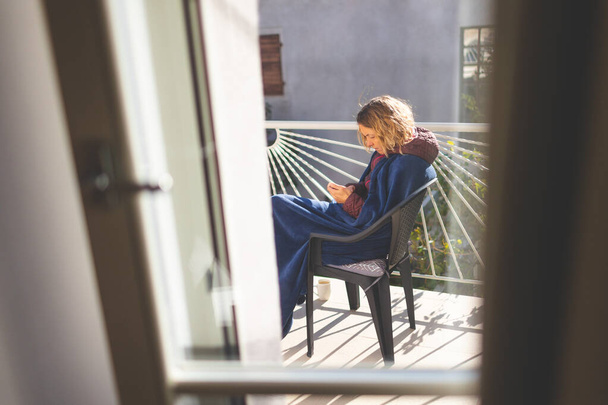 een meisje met een telefoon zit op een zonnig balkon, bedekt met een deken. - Foto, afbeelding