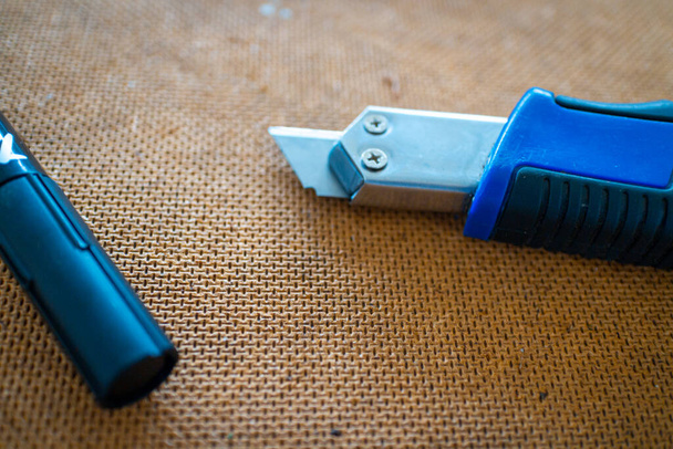 cuchillo de corte con mango azul y bolígrafo de marcado acostado en una superficie de trabajo de madera con enfoque en la placa de madera - Foto, Imagen
