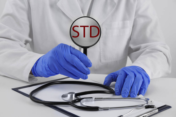 Doctor holding magnifying glass with abbreviation STD at white table, closeup - Foto, immagini