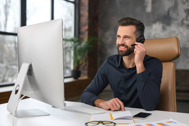 Office employees concept. Focused caucasian man wearing wireless headset is using laptop for online video conversation, talking with customers or coworkers - Φωτογραφία, εικόνα