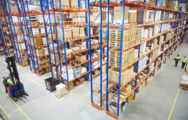 Blur warehouse background. Above view of warehouse workers moving goods and counting stock in aisle between rows of tall shelves full of packed boxes - Photo, image