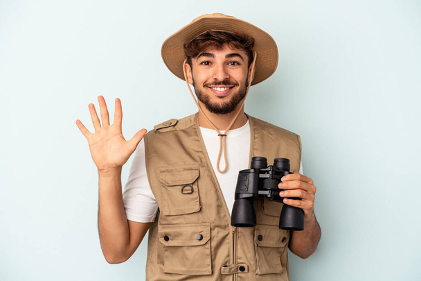 Young mixed race man holding binoculars isolated on blue background smiling cheerful showing number five with fingers. - 写真・画像