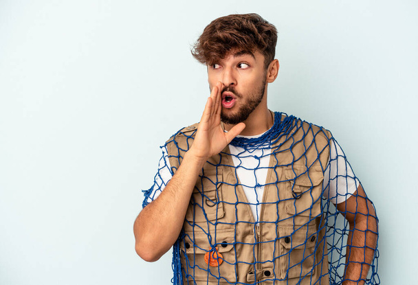 Young mixed race fisherman holding a net isolated on blue background is saying a secret hot braking news and looking aside - Fotó, kép