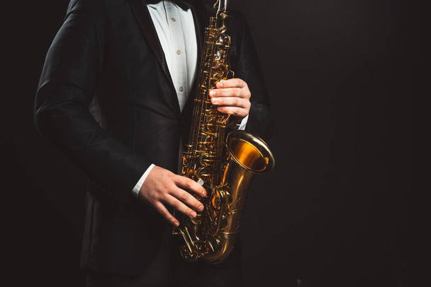 Saxophonist Plays Jazz. Male Musician in a Formal Black Suit Holds a Tenor Saxophone on a Dark Background. Saxophone Close-up. Copy space - 写真・画像