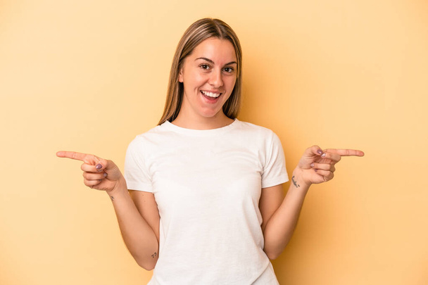 Young caucasian woman isolated on yellow background pointing to different copy spaces, choosing one of them, showing with finger. - Foto, Imagem