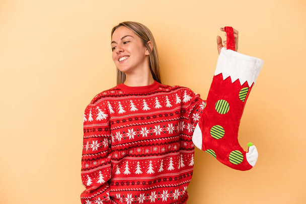 Young caucasian woman holding a elf sock isolated on yellow background looks aside smiling, cheerful and pleasant. - Photo, Image