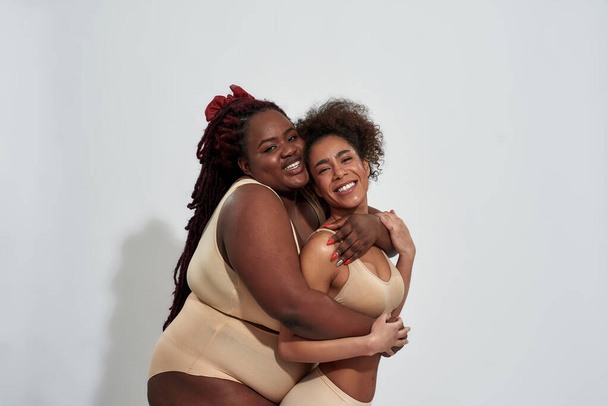Δύο χαρούμενες, λεπτές και υπέρβαρες Αφροαμερικανίδες με εσώρουχα αγκαλιασμένες και χαμογελαστές ενώ ποζάρουν μαζί απομονωμένες σε γκρι φόντο - Φωτογραφία, εικόνα