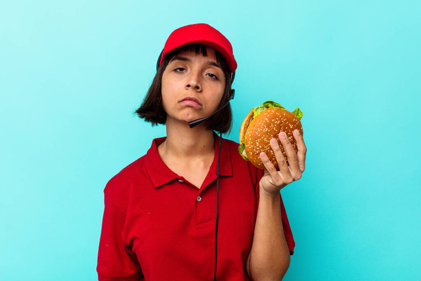 Joven mujer de raza mixta trabajadora de restaurantes de comida rápida sosteniendo una hamburguesa aislada sobre fondo azul - Foto, Imagen