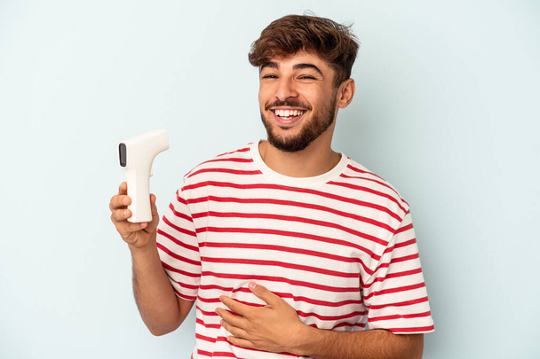 Young mixed race man holding thermometer isolated on blue background laughing and having fun. - Fotó, kép