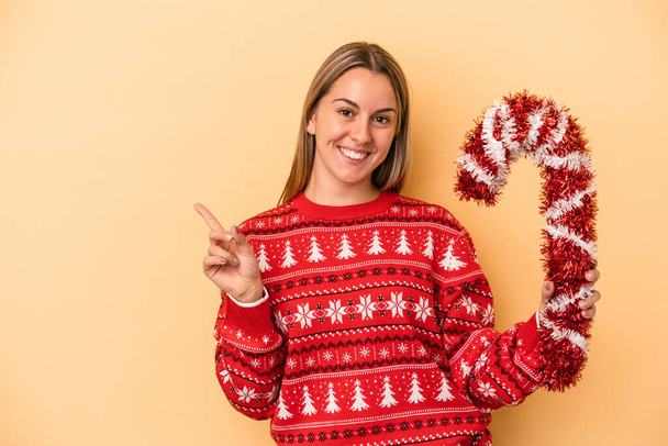 Young caucasian woman holding big christmas stick isolated on yellow background smiling and pointing aside, showing something at blank space. - Photo, Image