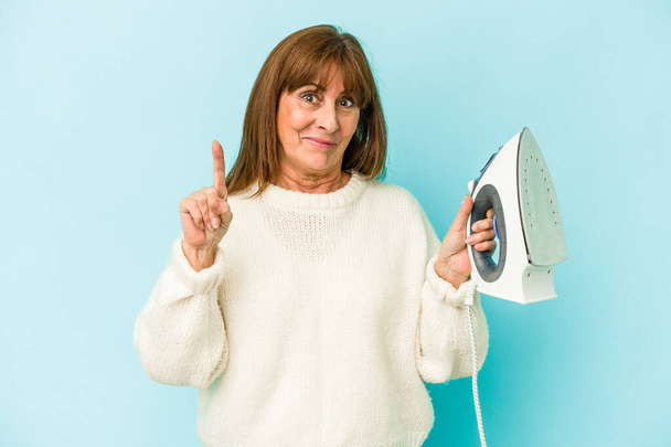 Middle age caucasian woman holding a iron isolated on blue background showing number one with finger. - Foto, Bild