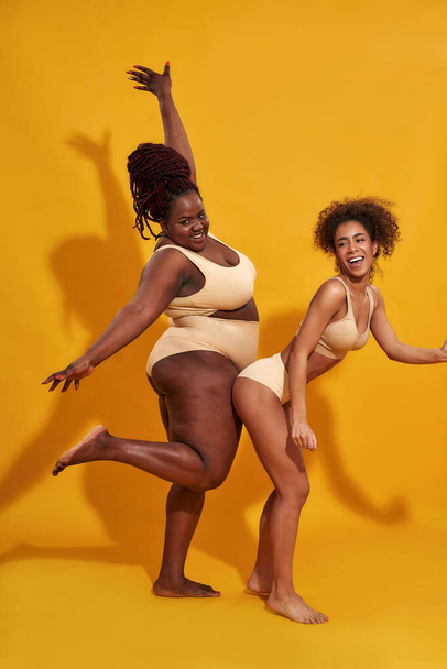 Deux excitées femmes africaines américaines minces et de taille plus en sous-vêtements beige s'amusant tout en posant ensemble isolé sur fond jaune - Photo, image