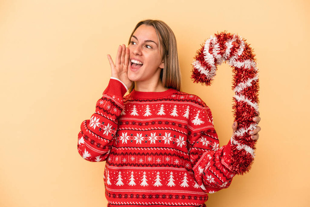 Joven mujer caucásica sosteniendo gran palo de Navidad aislado sobre fondo amarillo gritando y sosteniendo la palma cerca de la boca abierta. - Foto, imagen