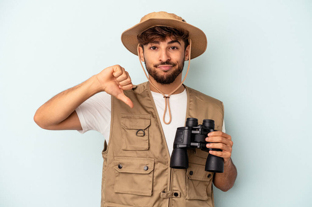 Young mixed race man holding binoculars isolated on blue background showing a dislike gesture, thumbs down. Disagreement concept. - 写真・画像