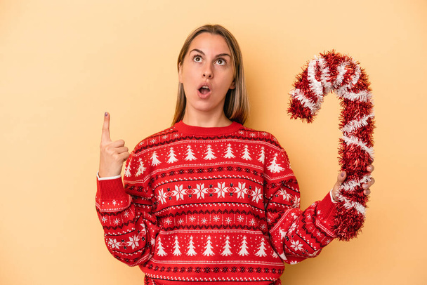 Young caucasian woman holding big christmas stick isolated on yellow background pointing upside with opened mouth. - Photo, Image