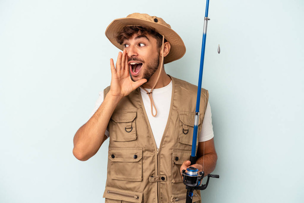 Young mixed race fisherman holding a rod isolated on blue background shouting and holding palm near opened mouth. - Photo, Image