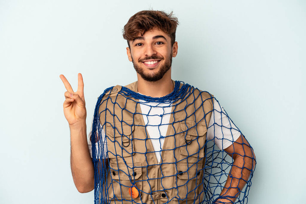 Young mixed race fisherman holding a net isolated on blue background joyful and carefree showing a peace symbol with fingers. - Fotó, kép