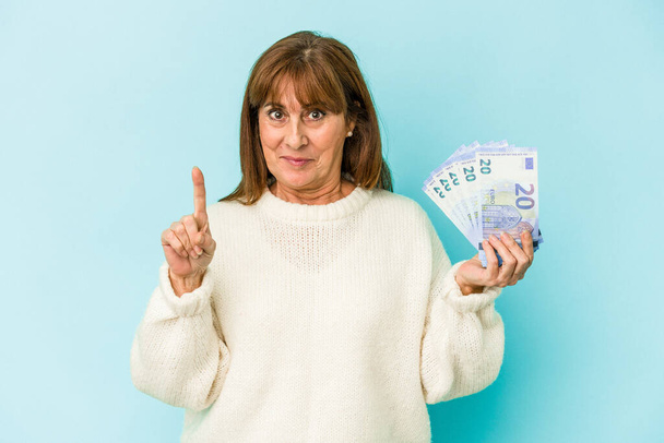 Середньовічна біла жінка тримає банкноти ізольовані на синьому фоні, показуючи номер один пальцем
. - Фото, зображення