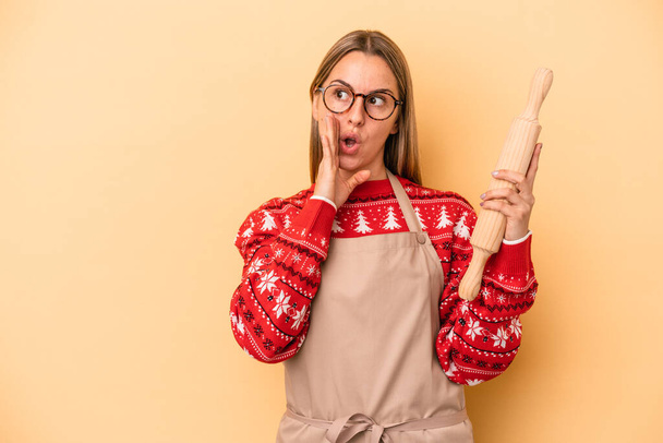 Junge Bäckerin macht Plätzchen für Weihnachten isoliert auf gelbem Hintergrund sagt eine geheime heiße Bremse Nachrichten und Blick zur Seite - Foto, Bild