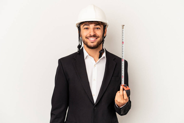 Jovem misto arquiteto homem vestindo um capacete e segurando medidor isolado no fundo cinza feliz, sorridente e alegre. - Foto, Imagem