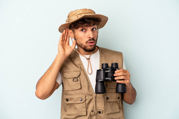 Joven hombre de raza mixta sosteniendo prismáticos aislados sobre fondo azul tratando de escuchar un chisme. - Foto, Imagen